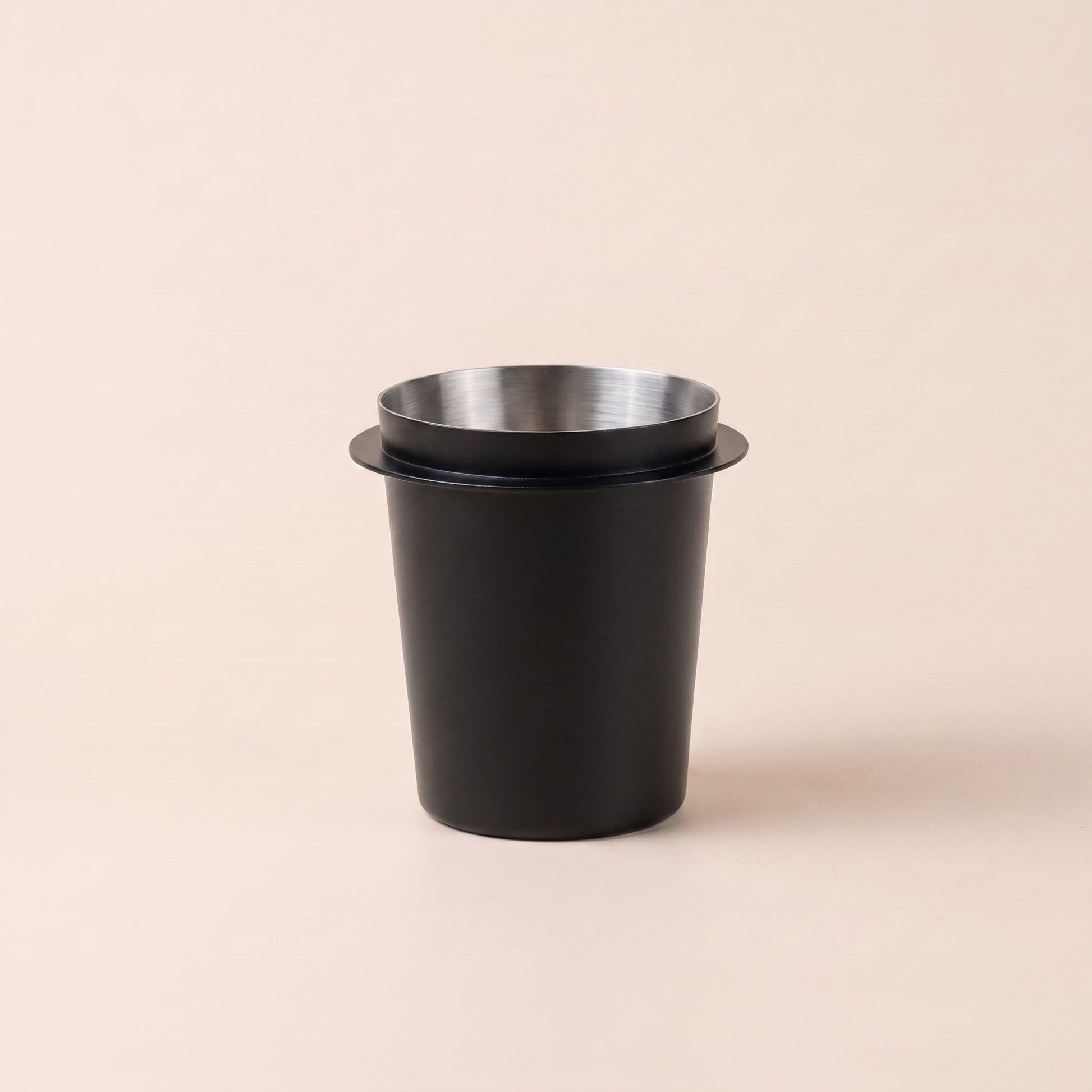 Black stainless steel espresso dosing cup, front three-quarter view on a warm cream background