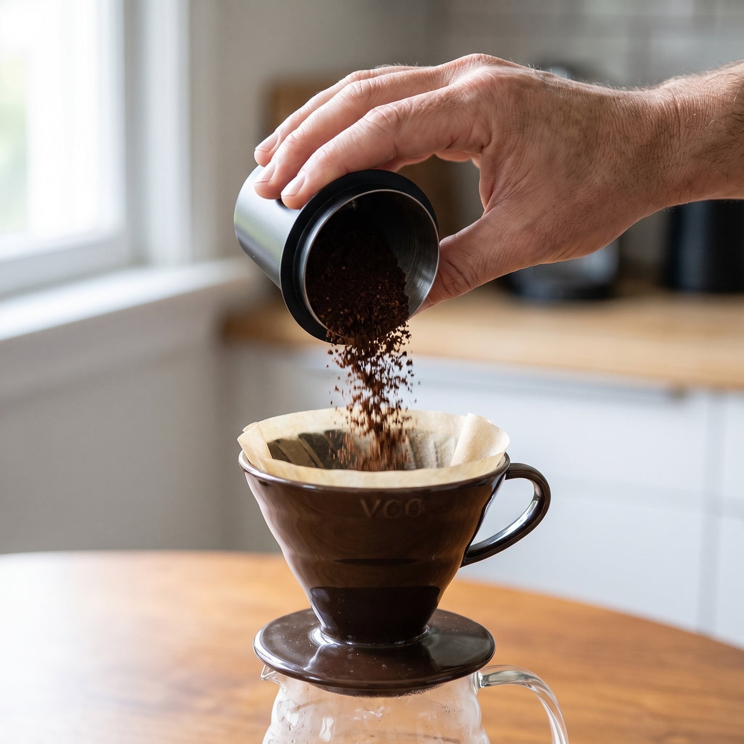 Hand pouring ground coffee from the dosing cup into a pour-over dripper on a home bench